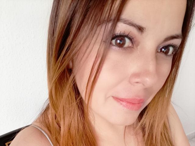Seher has light brown, straight hair with subtle highlights, brown eyes, and pink lips, smiling slightly. Her bare shoulders are visible, and she wears a thin strap.