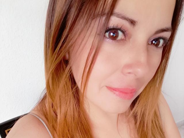 O rosto de Seher, com olhos castanhos, lábios rosados e cabelos castanho-claros com pontas loiras, vestindo uma blusa com alças finas, olhando ligeiramente para a direita com um sorriso discreto..