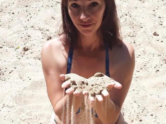 MissieLorrie, com cabelo castanho, usando um biquíni azul, segurando areia nas mãos, sorrindo, em uma praia ensolarada, seus pequenos seios e mamilos levemente visíveis através da areia..