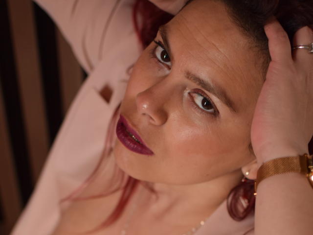 SalmaBrown heeft donkere ogen, rode lippenstift en een lichtrode huid, met een hand in haar roze haar, draagt een roze top en een gouden polshorloge, en kijkt omhoog..