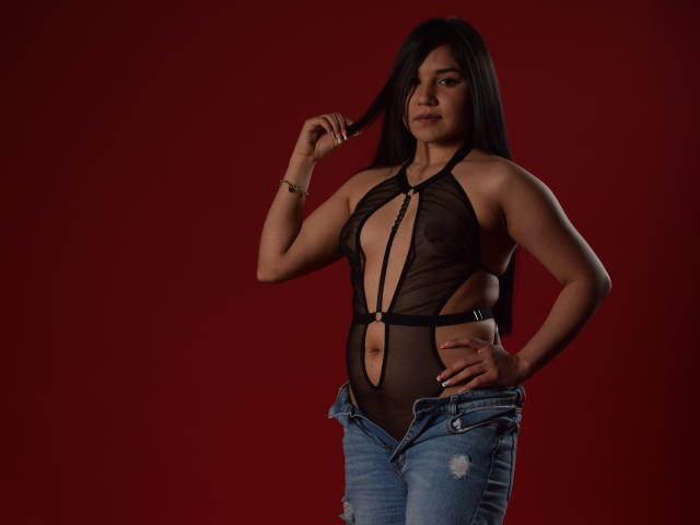 Melissa Blaiir indossa un top di lingerie nero trasparente con spalline, che mette in mostra i suoi seni, con un taglio profondo che rivela i capezzoli, e jeans blu sbottonati, su uno sfondo rosso..