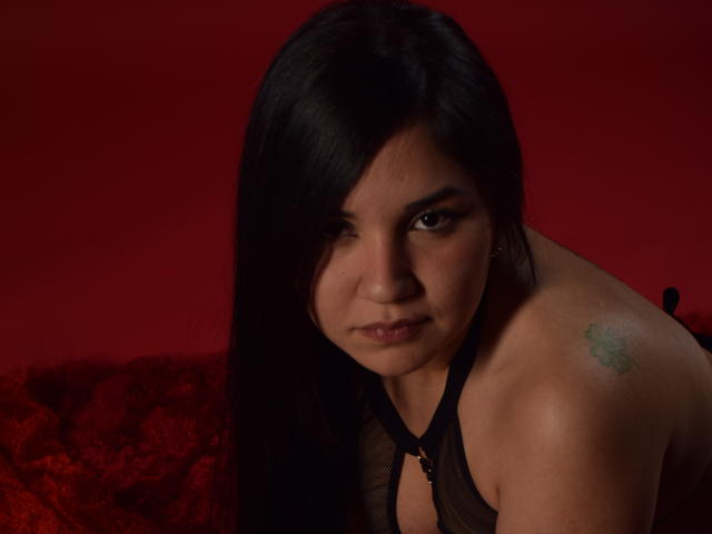MelissaBlaiir a de longs cheveux noirs, une expression sérieuse et un petit tatouage vert sur son épaule droite. Elle porte un haut en maille noire qui laisse apparaître son dos nu..
