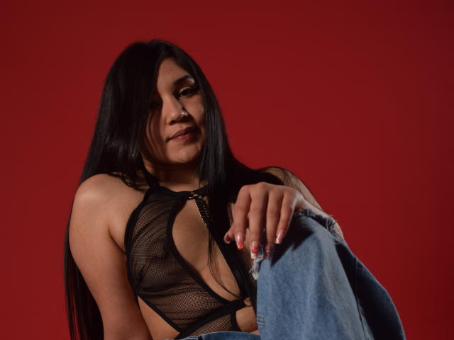 MelissaBlaiir, avec de longs cheveux noirs, portant un haut noir transparent qui révèle ses seins, un jean bleu et un collier en or, assise devant un fond rouge, regardant vers le bas..