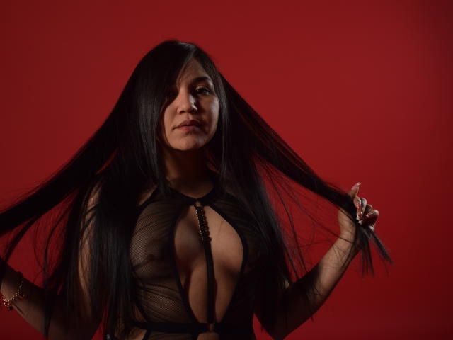 MelissaBlaiir, avec de longs cheveux noirs, les tenant, portant un haut noir transparent qui expose ses seins, sur un fond rouge, regardant l'appareil photo avec un léger sourire..
