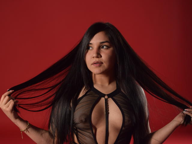 Melissa Blaiir, con lunghi capelli neri raccolti, indossa una trasparente maglia nera che rivela il seno e un dettaglio a catena, su uno sfondo rosso, guardando leggermente di lato..