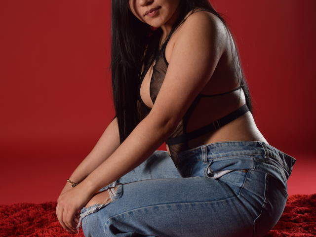 MelissaBlaiir with long black hair, wearing a sheer black bra that exposes her tits and blue jeans, kneeling on a red textured surface, looking down with a slight smile.