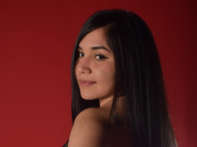 Le visage de MelissaBlaiir, regardant par-dessus son épaule avec de longs cheveux noirs, une peau brun clair et un léger sourire, montrant ses petits seins mignons sur un fond rouge uni..