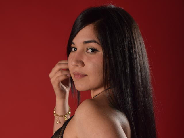MelissaBlaiir tiene el pelo largo y negro, una sonrisa sutil, y está mirando por encima del hombro, con una pulsera dorada en su muñeca derecha. Sus senos son parcialmente visibles..
