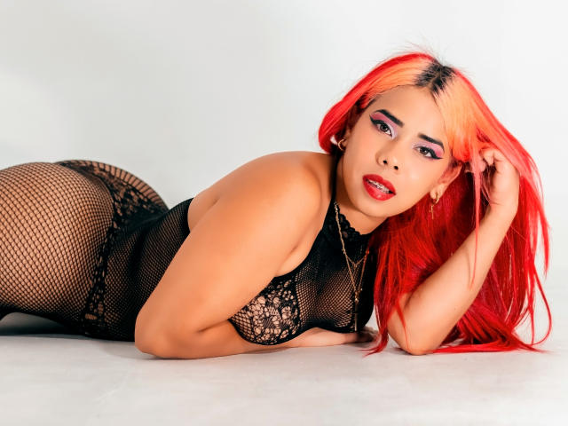Valentina, con cabello rojo, acostada de lado, vistiendo lencería de malla negra, mostrando sus pechos, trasero y vulva, mirando a la cámara con lápiz labial rojo..