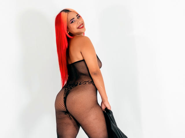 ValentinaCute with bright red hair, smiling, wearing a black fishnet bodysuit, showing her ass, holding black fabric, looking over her shoulder.