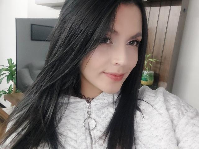 LinaStars has long black hair, pale skin, and is wearing a white textured shirt with a silver necklace. She's smiling softly, showing subtle pink lips.