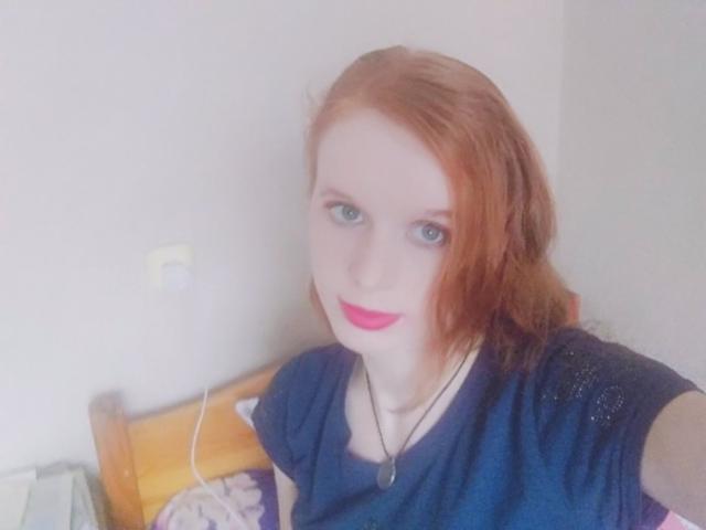StaceyLove has red hair, blue eyes, and pink lipstick. She's wearing a blue shirt with a black necklace. Her tits are partially visible. Her ass is not shown.