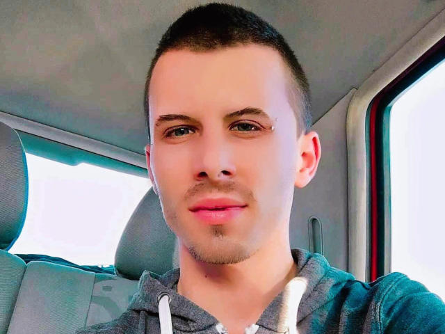 TwinkMario has short dark hair, green eyes, and a slight mustache. He's wearing a grey hoodie with white drawstrings, looking at the camera with a subtle smile.