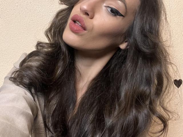 VievienneClemente has long, wavy brown hair, pinkish lips, and black winged eyeliner. She's wearing a beige top, looking up with a slight pout. Her ass is not visible.