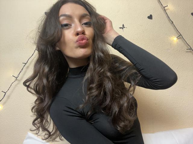 Vievienne Clemente, avec ses longs cheveux bruns ondulés, ses lèvres pulpeuses et son haut noir à manches longues, touchant ses cheveux, avec un maquillage discret, sur une photo d'elle-même..