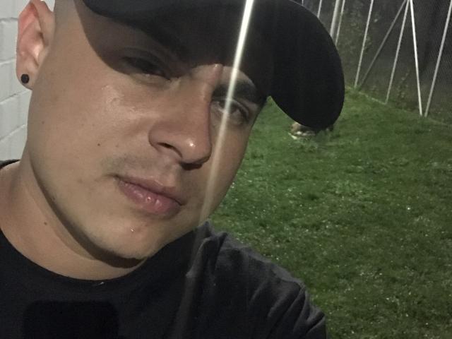 BenSuarez, con un cappello nero, un naso leggermente lucido, una camicia nera e un foro nell'orecchio nero, in piedi sull'erba verde, che mostra un sorriso appena accennato. Luce intensa sul viso..