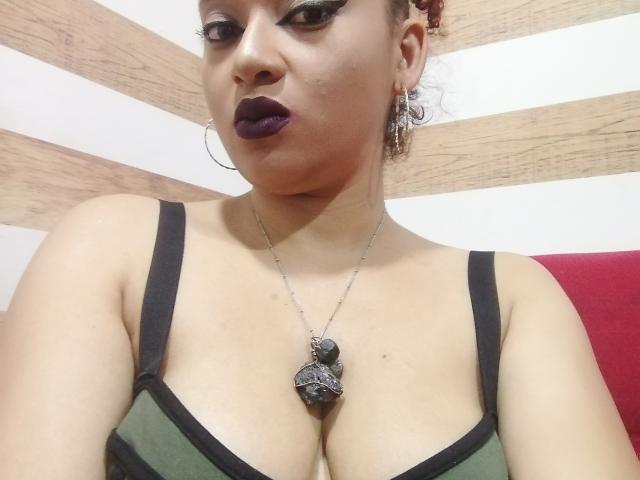 VioletaSwitch with dark purple lipstick, hoop earrings, and a necklace with a decorative pendant, wearing a green and black bra, showing her large tits and a confident expression.