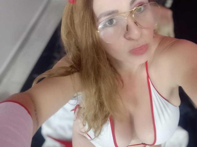 PaulinaMoon con gli occhiali, rossetto rosa e capelli rossi, indossa un abito bianco e rosso con un profondo scollo, che mostra il seno. Sta guardando verso l'alto, con le labbra tumefatte..