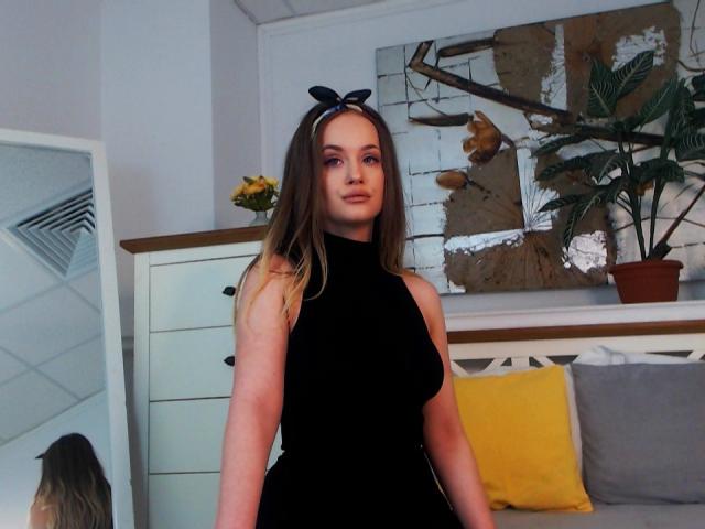 SophieXDee, con cabello largo y castaño, un vestido negro sin mangas y una diadema con lazo negro, está de pie en una habitación, mostrando su trasero y sus pequeños senos, con un cojín amarillo en el fondo..