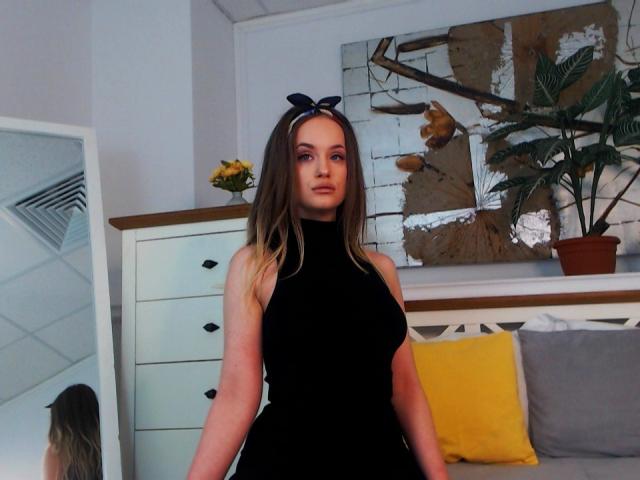 SophieXDee heeft lang bruin haar, een zwart mouwloos jurkje met een hoge hals, en een zwarte strik in haar haar. Haar achterkant is bedekt, en ze staat in een kamer met gele en grijze kussens..