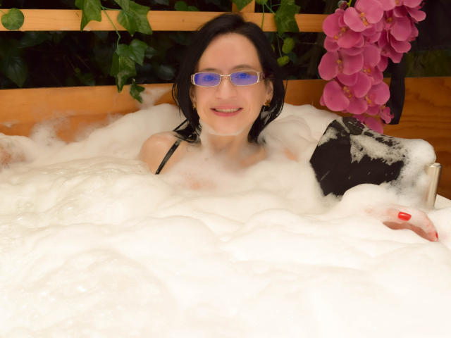 AlanaMoneth with blue-tinted glasses, black hair, and red nails, sitting in a bubbling hot tub, partially covered in white foam, holding a black bag, with pink flowers.