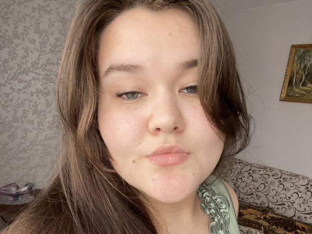 De volle, lichtroze lippen van IsadoraStefanie zijn lichtjes samengeknepen in een kusgezicht, met haar groene ogen half open en haar bruine haar dat over haar gezicht valt. Ze heeft een kleine moedervlek..