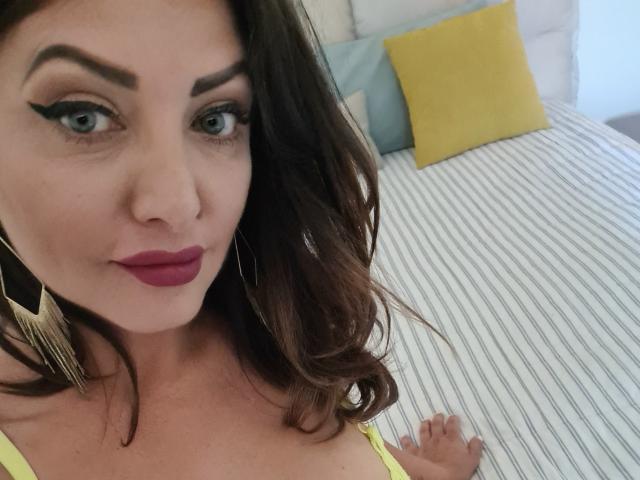 Miky69I has blue eyes, dark brown wavy hair, and red lipstick. She's wearing a yellow top, showing her tits, and large gold earrings. Her right hand rests on a bed.