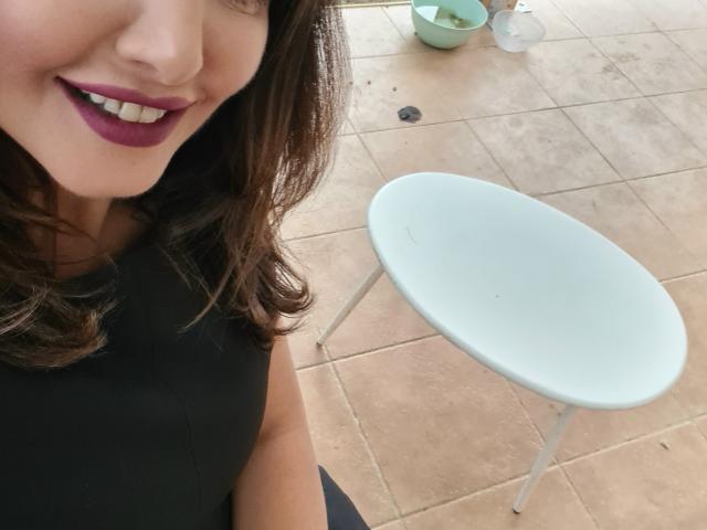 Il viso di Miky69I con rossetto viola scuro, che sorride, indossa una canotta nera, ha capelli castani ondulati, una sedia ovale bianca con gambe in metallo e un pavimento piastrellato con una ciotola verde..