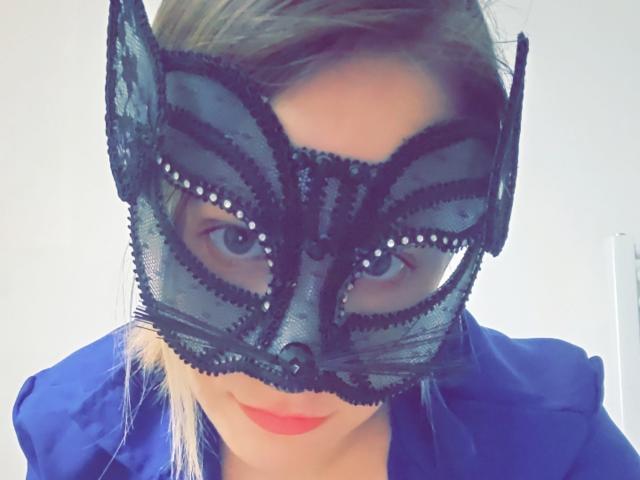 DreamSha wearing a black lace Batman mask with bead detailing, blue shirt, and red lipstick, showing off her tits slightly visible beneath the mask.