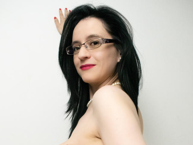 AlanaMoneth with black hair, glasses, red lipstick, pearl necklace, and red nail polish, smiling, showing her bare shoulders and back, hand on wall.