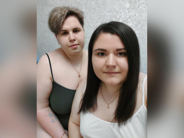 BeckyAndEllen with short, curly brown hair and small tits, wearing a green tank top, sits behind BeckyAndEllen with long, black hair and small tits, in a white tank top.