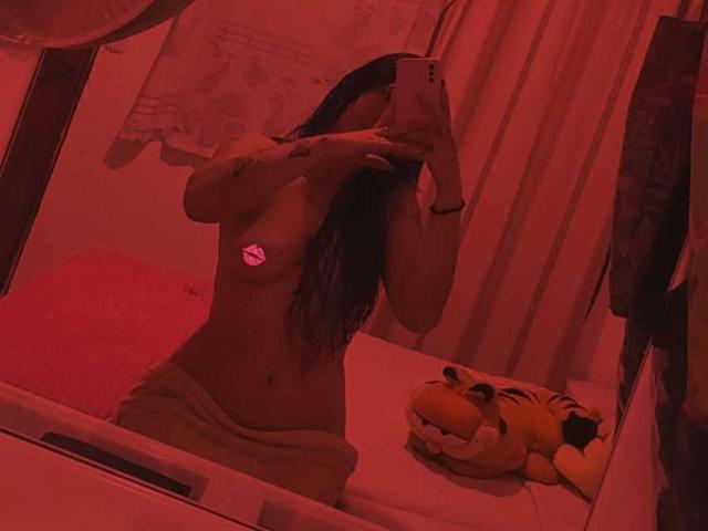 Selfie no espelho com luz vermelha de LexySmithX, sem blusa, com um adesivo rosa cobrindo os seios, cabelo longo e escuro, segurando o celular, toalha marrom enrolada na bunda, tigre de pelúcia no balcão..