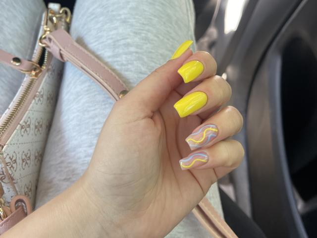 La mano di SugarrX, con unghie di un giallo brillante e un'unghia rosa con un motivo sull'anulare, che tiene una borsa rosa, in contrasto con i pantaloni grigi, all'interno di un'auto..