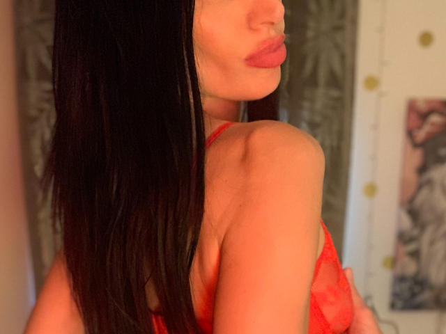 AlicianeFrancaise con lunghi capelli neri, reggiseno di pizzo rosso, labbra rosa carnose e una spalla scoperta, che mette in mostra il seno. Una luce calda esalta la sua pelle..
