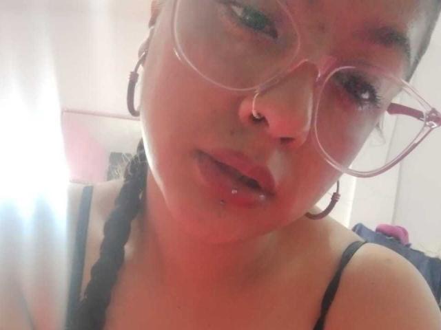 Brunetta Lia, con occhiali rosa, piercing al naso e capelli intrecciati, mostra le sue labbra leggermente aperte, bagnate e lucide, indossa un top nero con spalline, in un selfie luminoso e leggermente sfocato..