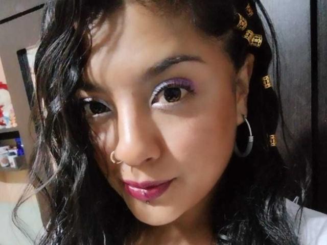 BrunetteLiaSex with dark wavy hair, purple eye makeup, hoop earrings, nose ring, pink lipstick, and a lower lip piercing, looking slightly to the side with a subtle smile.
