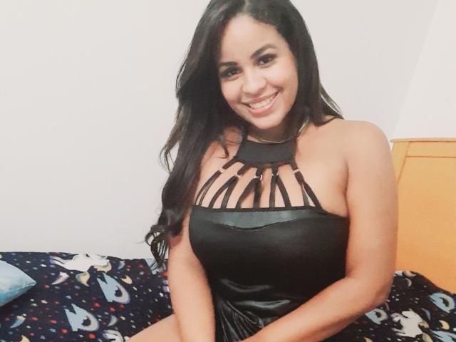 Adannia has long black hair, smiling, wearing a black strapless dress with cut-out chest, showing her tits, sitting on a bed with a blue, patterned blanket.