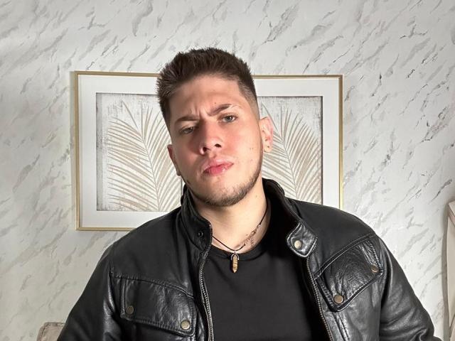 SebastiianMaxime, com cabelo castanho curto, pele clara, jaqueta de couro preta, camisa preta, colar, expressão emburrada, em pé em frente a uma obra de arte com folhas emolduradas..