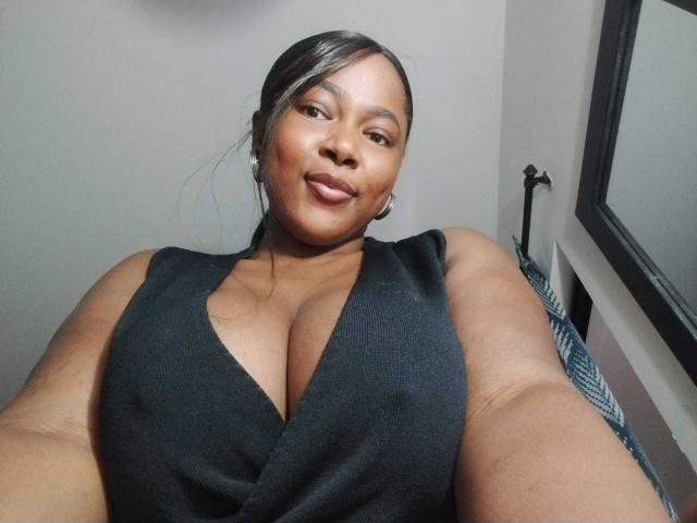 Emira, atractiva, con grandes senos, vestida con una blusa negra de cuello en V pronunciado, sonriendo a la cámara, con aretes blancos, cabello oscuro en una trenza lateral, tomando un selfie..
