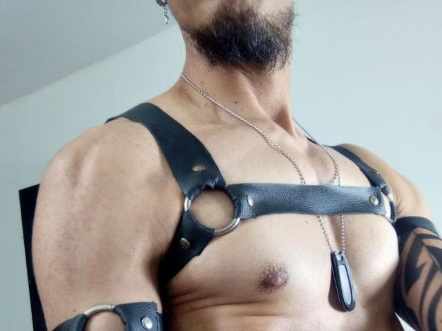 JeikkoHot con un'imbracatura in pelle nera, medagliette e barba, mostra un petto muscoloso e scoperto con piccoli capezzoli, e un tatuaggio sul braccio destro..