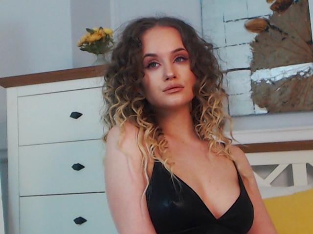 SophieXDee with curly blonde hair, wearing a black v-neck, showing her tits, standing in a room with a white dresser and yellow flowers in the background.