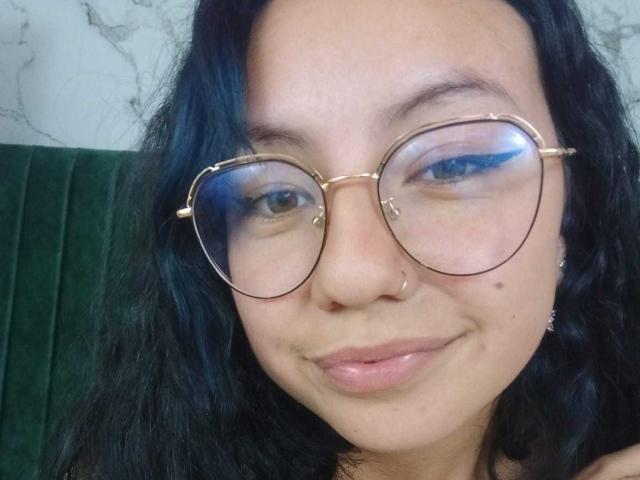 VeroHotLove con occhiali rotondi, eyeliner blu, capelli neri ondulati, un anello al naso e una piccola lentiggine sulla guancia destra, sorride dolcemente. Le sue natiche non sono visibili..