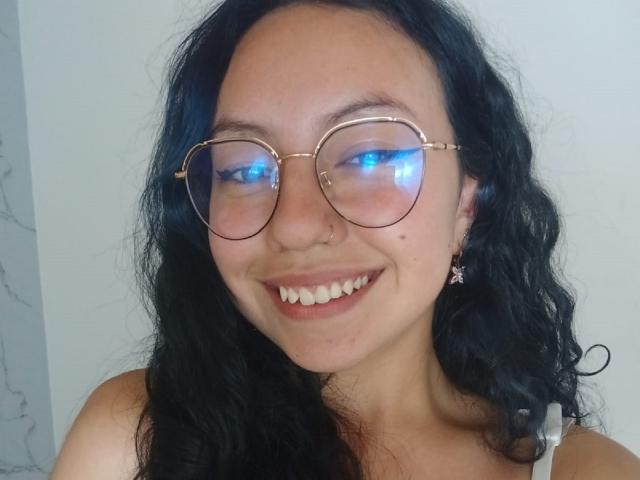 A VeroHotLove tem cabelos pretos cacheados, óculos com moldura dourada que refletem luz azul, um piercing no nariz e um sorriso que mostra dentes brancos. Ela está vestindo uma blusa branca com alças e brincos de estrela..