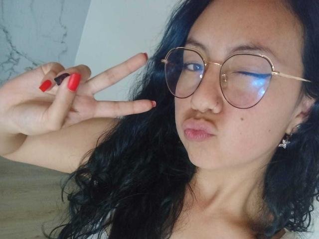VeroHotLove mit welligem, schwarzem Haar, roten Nägeln, runden Brillen und einem Nasenring, die ein Friedenszeichen macht, mit leichtem Schmollmund und einem kleinen Muttermal auf der Wange..