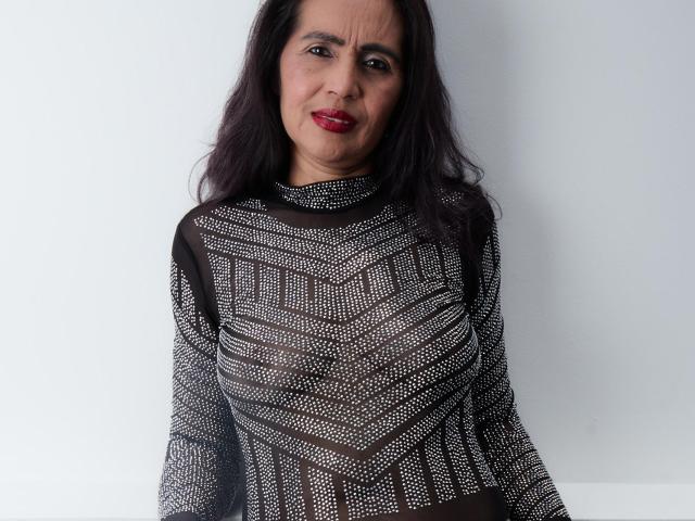 MoxiaHot with dark hair, red lipstick, and wearing a black, sheer, patterned top, showing her tits through the fabric. She's smiling slightly.