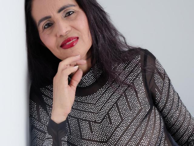 MoxiaHot has dark hair, red lipstick, and wears a black, patterned, see-through top with white dots. She's smiling, touching her chin with her right hand.