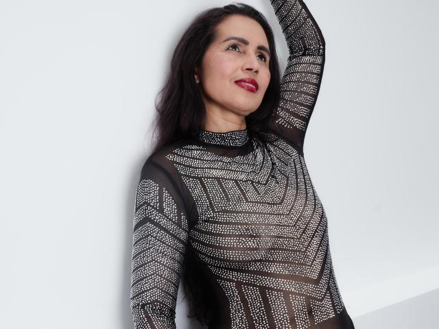 MoxiaHot with dark hair, red lipstick, wearing a black, sheer, patterned top, arm raised, smiling, white background, sexy, attractive, medium breasts, confident, stylish, standing, looking up.