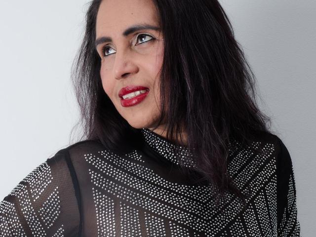 MoxiaHot with dark, wavy hair, red lipstick, and black sheer top with white polka dots, looking sideways with a slight smile. Boobs partially visible through the top.