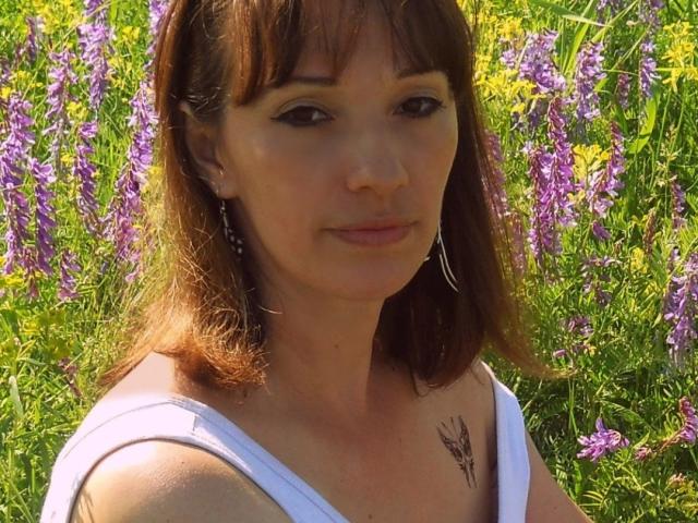 MissieLorrie has brown hair, a white top, and a small butterfly tattoo on her right shoulder. She's wearing earrings and looking at the camera with a slight smile.
