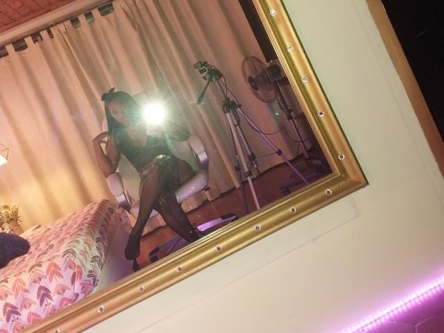 TiffanySammy in black lingerie, fishnet stockings, and two buns in her hair, taking a mirror selfie with a flash, showing her ass and legs, in a bedroom.